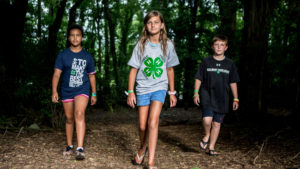Three children walking toward the camera side-by-side in the forest as part of an Extension 4-H program