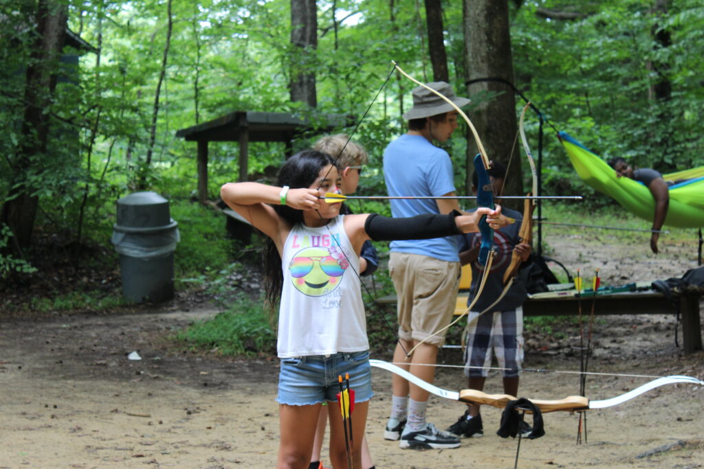 A female youth participating in archery with a bow and arrow. She has long hair, white tip and jean shorts.
